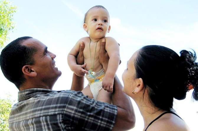 Padre sostiene al hijo en los brazos, mientras la madre los observa.